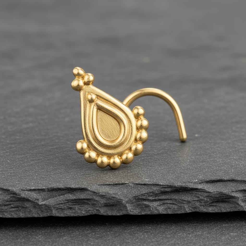 A gold-colored nose stud with a decorative design is shown against a gray background.