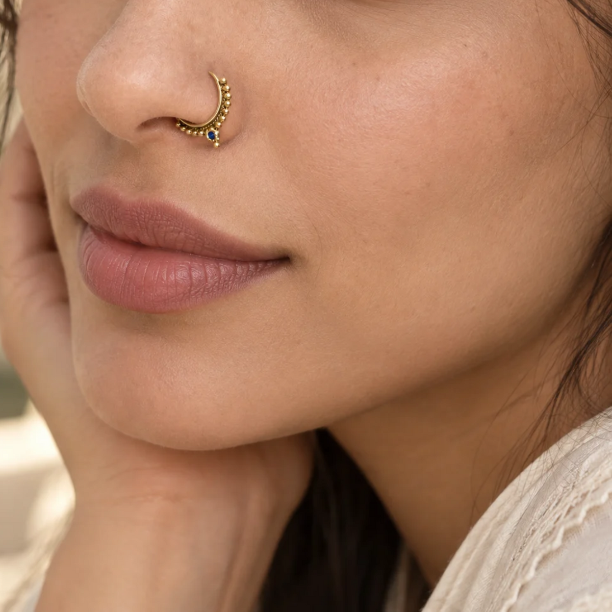A close-up of a woman's face, focusing on her nose and lips, with a gold nose ring and a delicate blue gemstone accent.