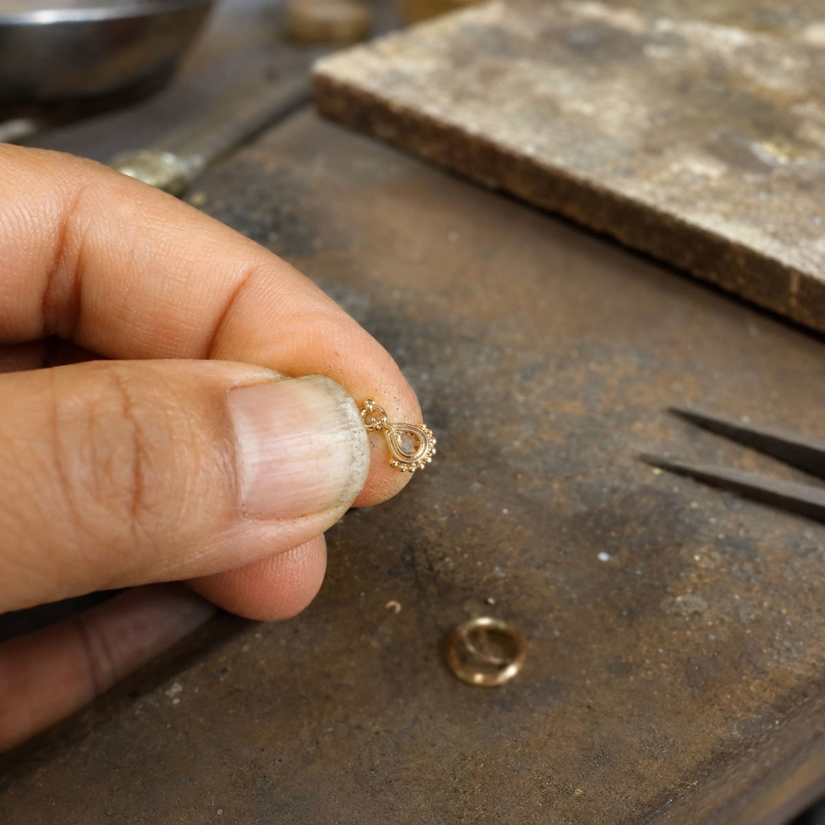 A person's hand holding a small gold-colored object, possibly a ring or a piece of jewelry, with a pair of pliers visible in the background.