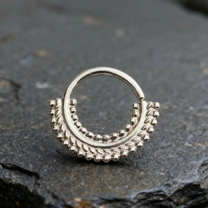 A silver-colored metal piercing ring with a decorative design on a dark surface.