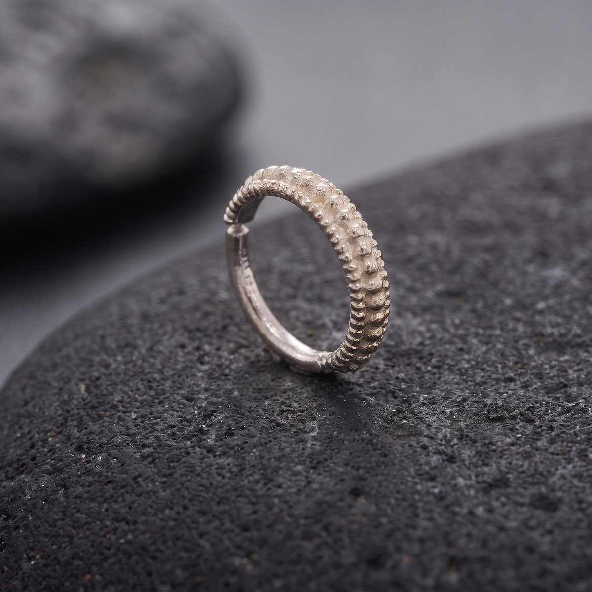A silver ring with a textured design sits on a dark surface, possibly a rock or a stone.