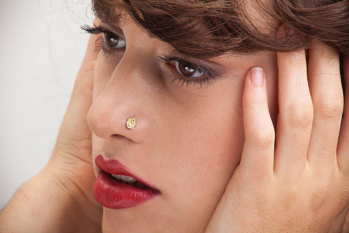A close-up of a woman's face, with her eyes closed and her hands covering her ears. She has red lipstick and a nose stud.