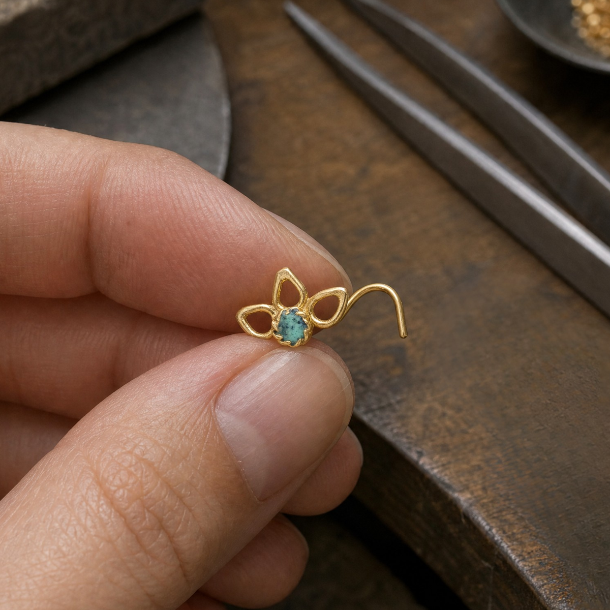 A hand holding a small gold-colored nose ring with a blue gemstone in the center.