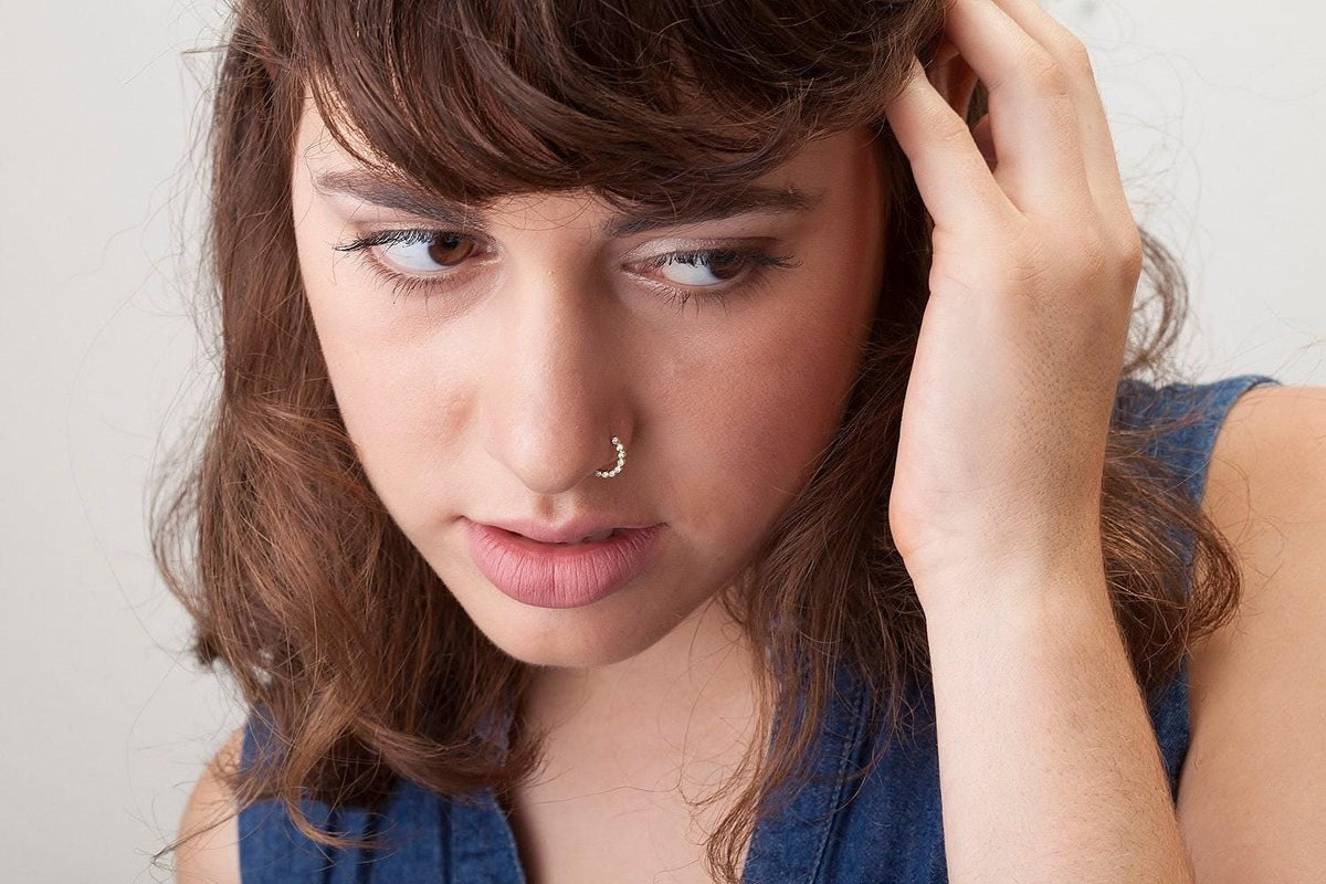 A close-up portrait of a young woman with brown hair, wearing a blue top and a nose ring, looking directly at the camera with a serious expression.