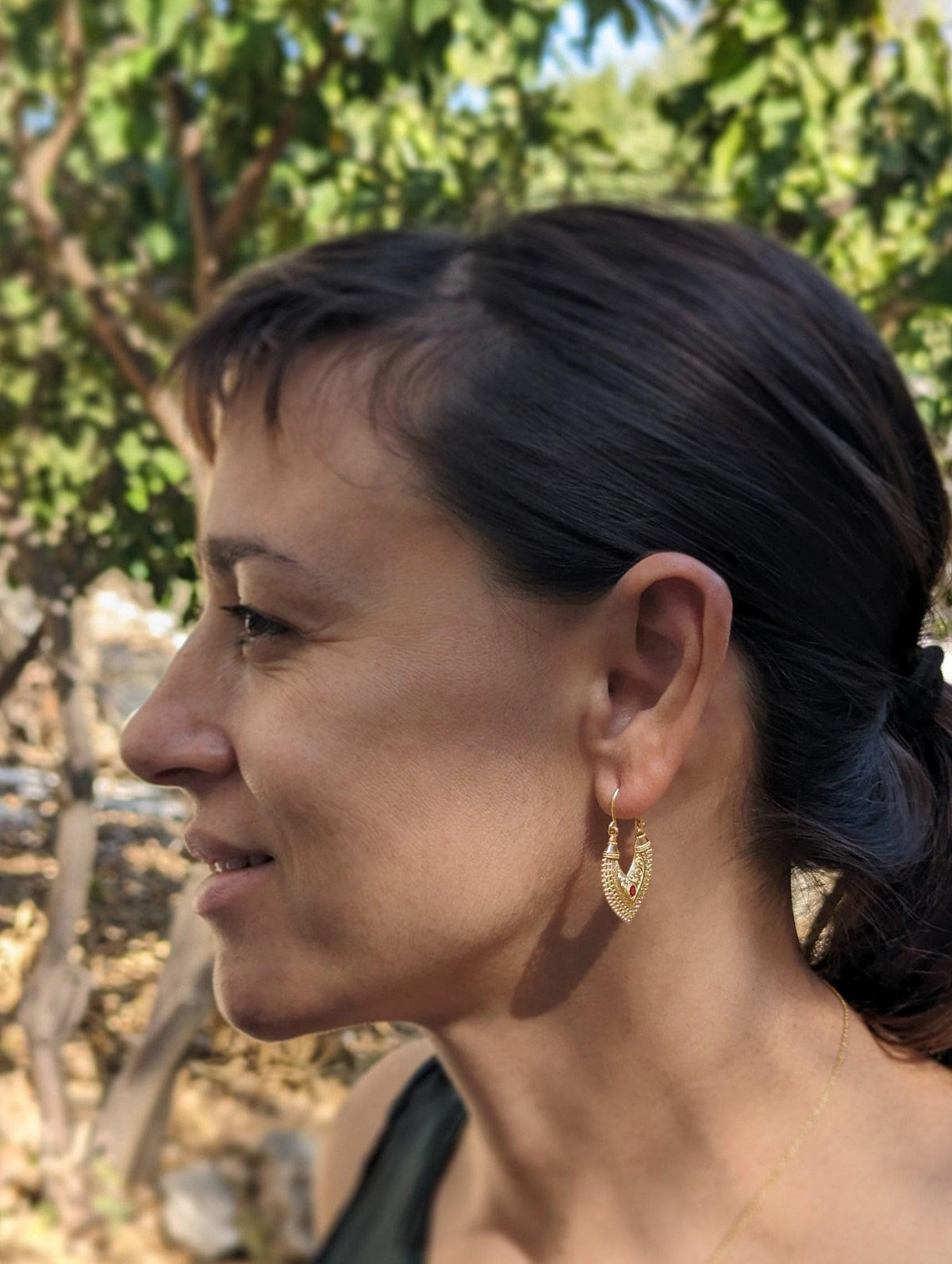 A woman with dark hair is smiling and wearing a pair of gold earrings with a heart-shaped design.