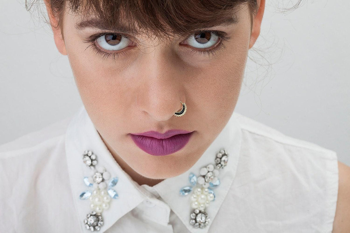 A close-up portrait of a woman with dark hair and striking blue eyes, wearing a white collared shirt with a decorative necklace.