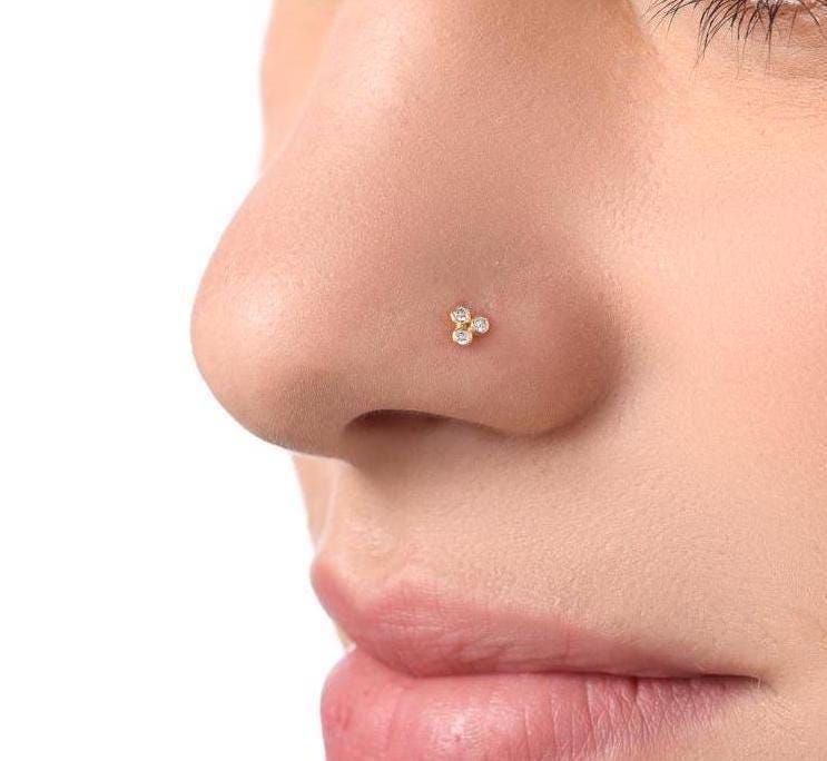 A close-up of a person's nose with a small gold-colored nose stud featuring three small diamonds.