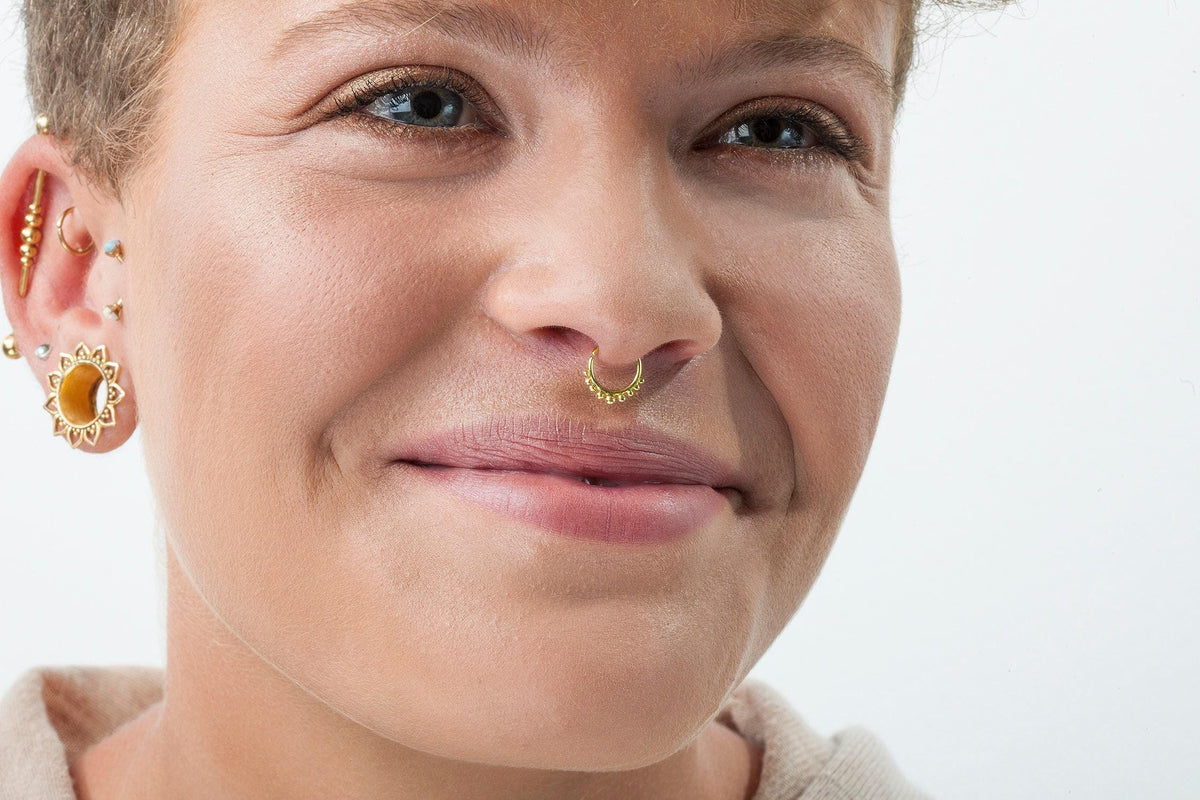 A close-up of a woman's face, with a nose ring and earrings visible. She has a warm, friendly expression on her face.