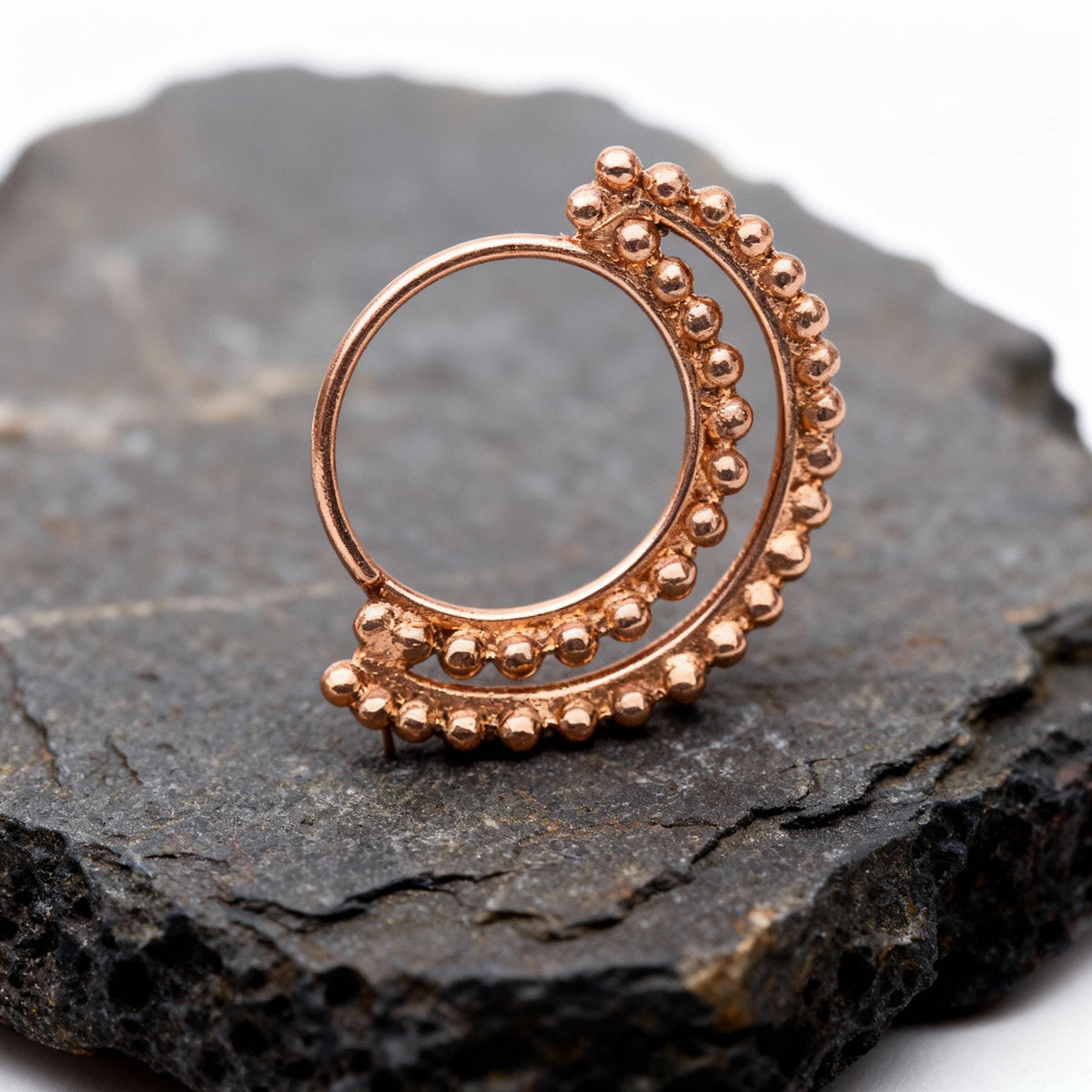 A copper-colored metal ring with a circular design and a row of small, round beads protruding from it, resting on a dark, textured surface.