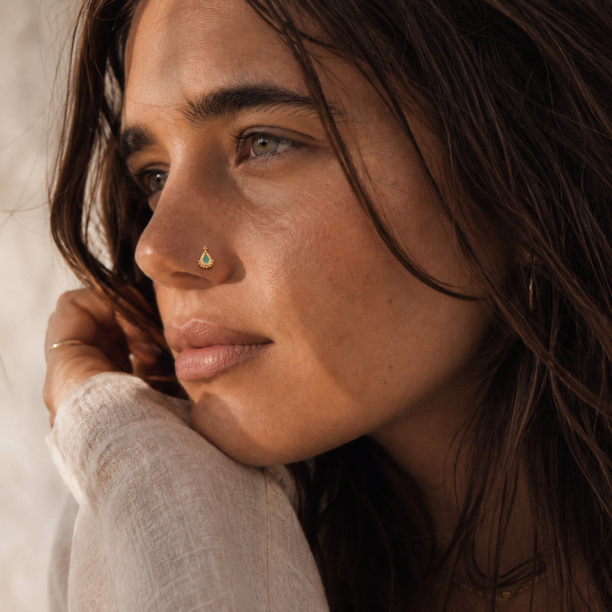 A close-up portrait of a woman with long, dark hair, wearing a white top and a nose ring.