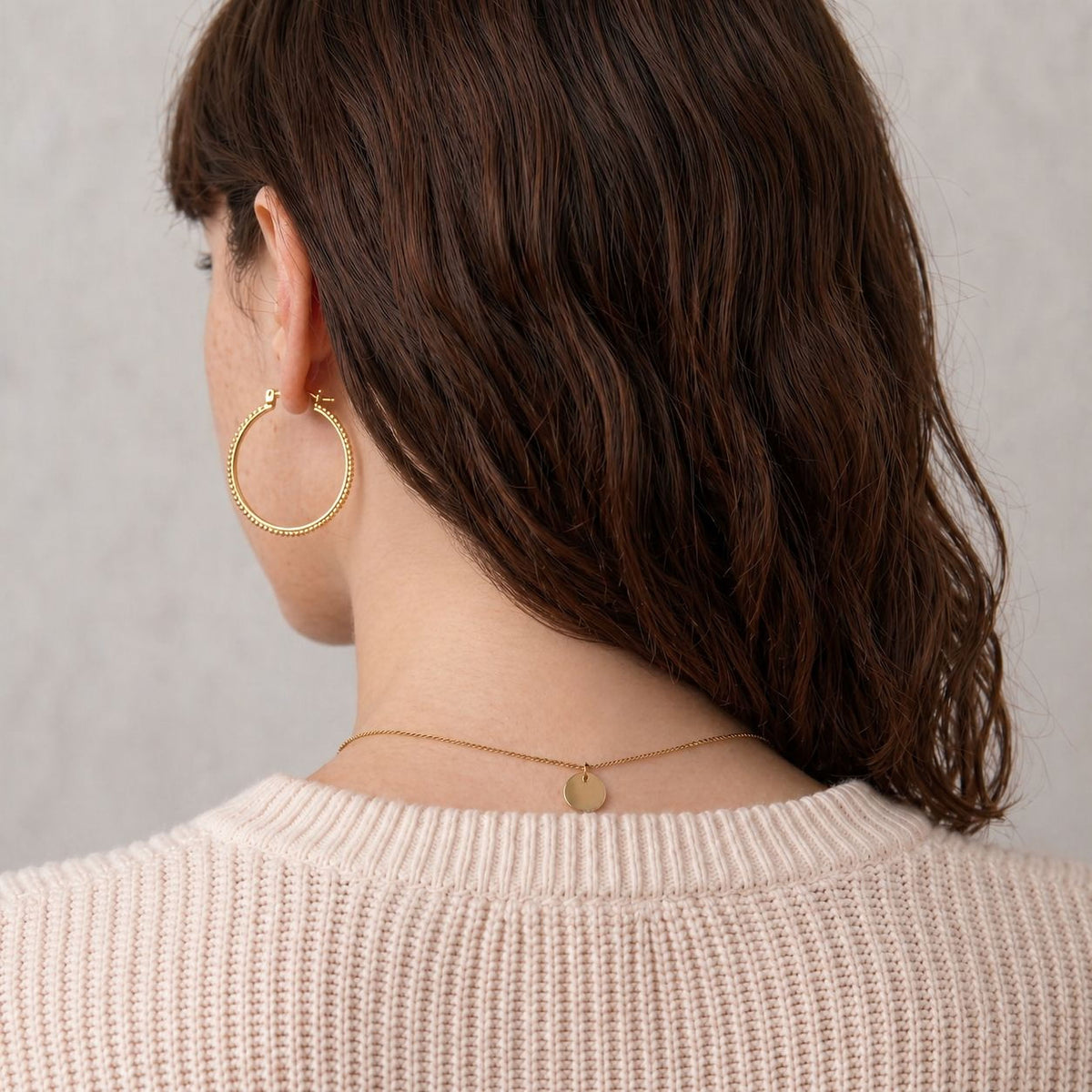 the back view of a woman with long brown hair wearing a white sweater and gold hoop earrings.
