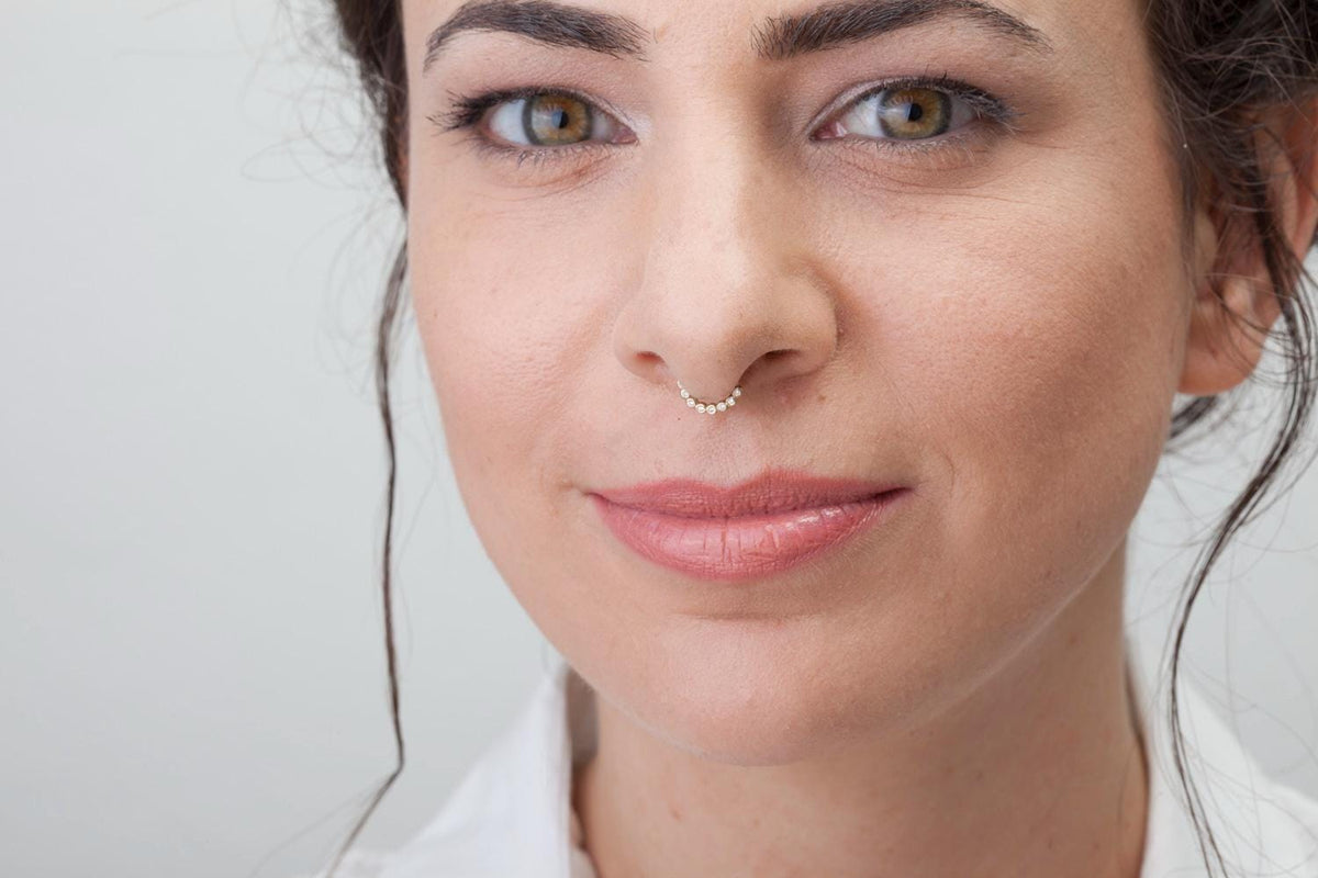 A close-up portrait of a woman with brown hair and a nose ring, wearing a white shirt.