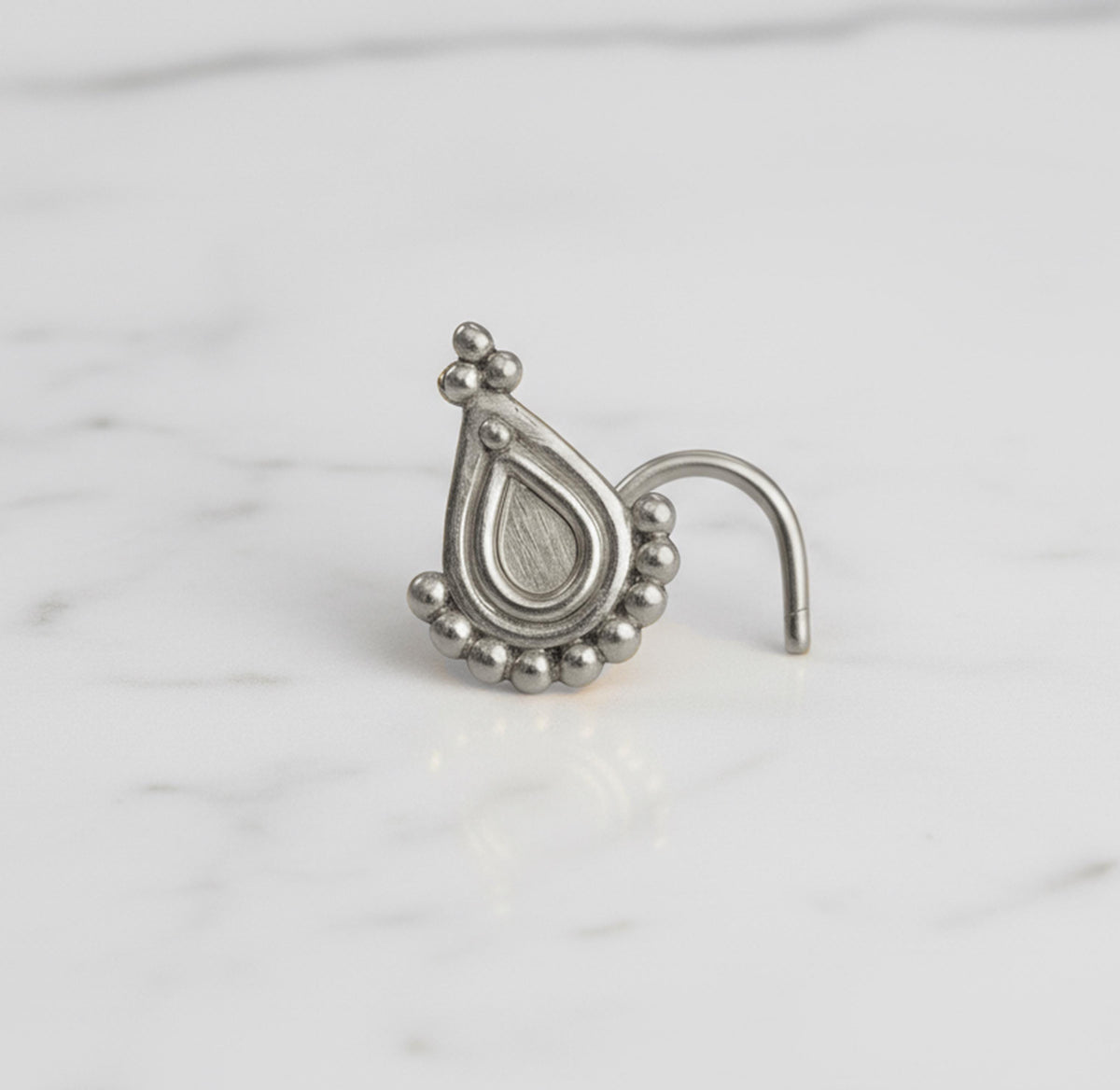 A silver nose ring with a decorative pendant is shown on a white marble surface.