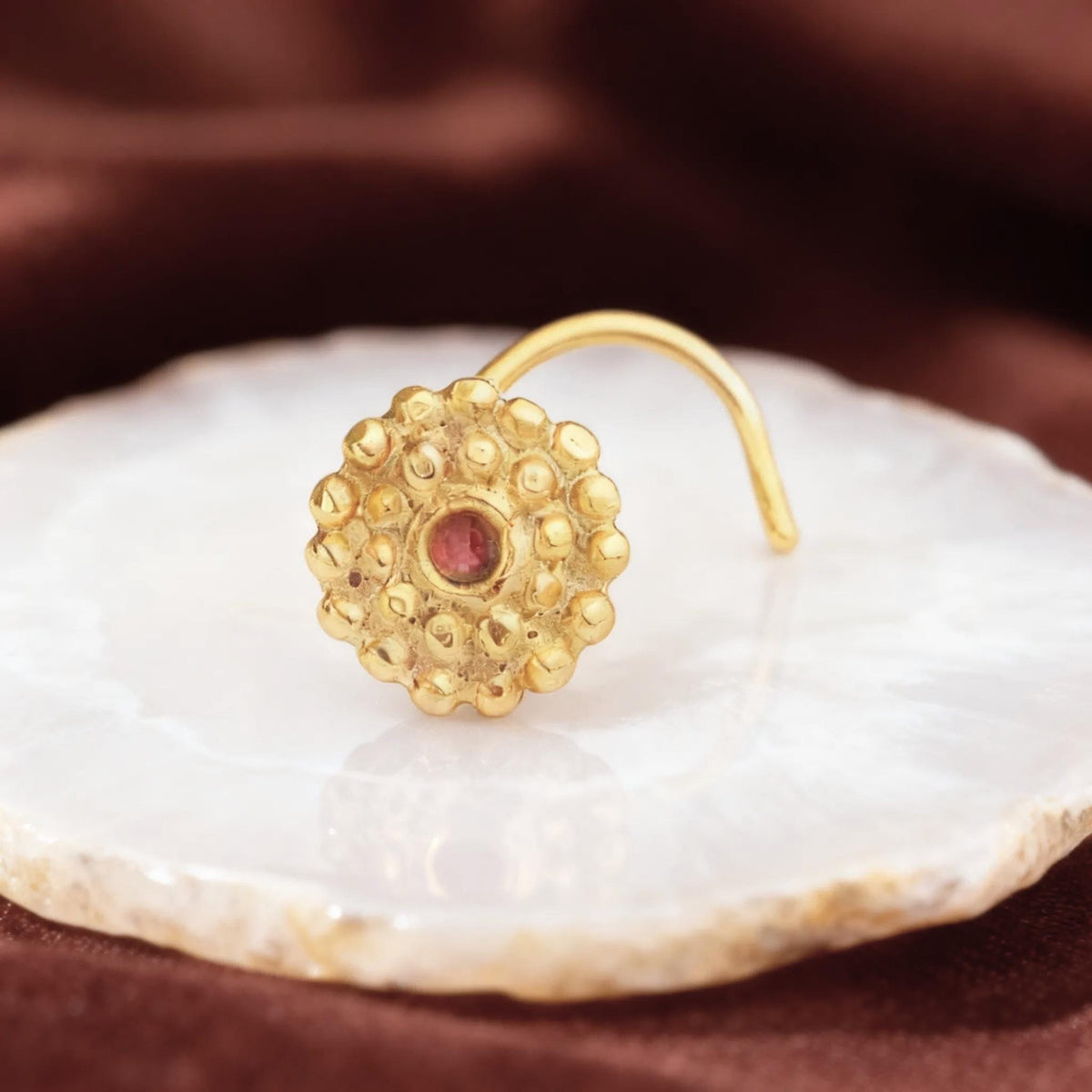 A gold-colored nose ring with a red gemstone is displayed on a white surface against a red background.