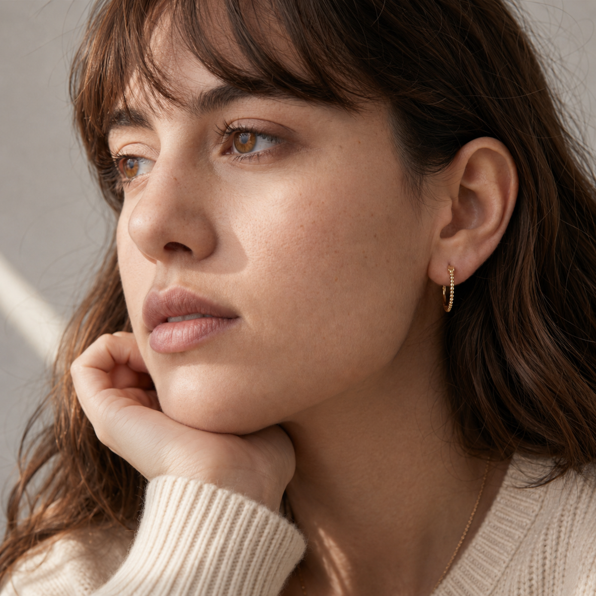 A close-up portrait of a woman with long brown hair, wearing a white sweater and earrings, looking pensively to the side.