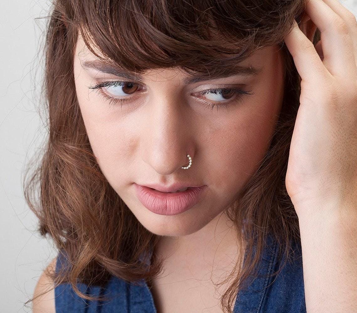 A close-up portrait of a young woman with brown hair, wearing a blue top and a nose ring, looking pensively at the camera.