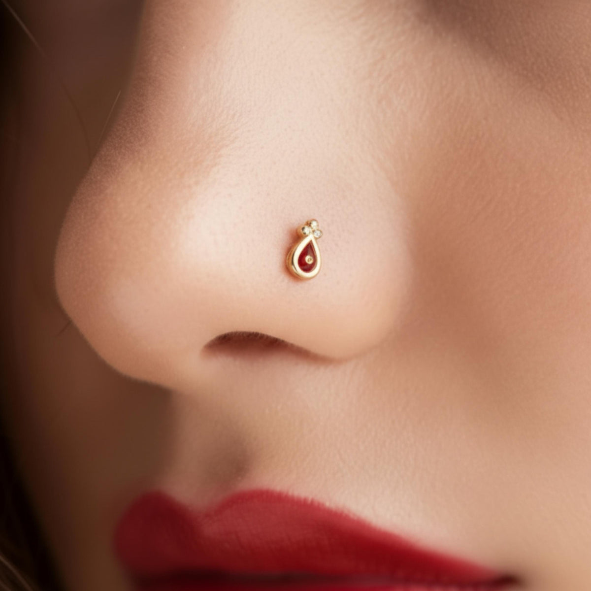 A close-up of a person's nose with a small, ornate gold nose ring featuring a red gemstone.