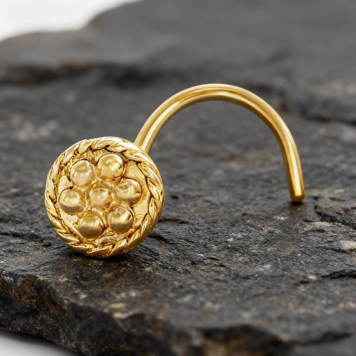 A gold-colored nose ring with a circular design is resting on a dark surface.