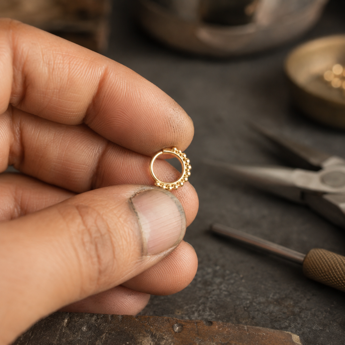 A hand holding a small gold-colored ring with a decorative element on it, surrounded by various jewelry-making tools and materials on a workbench.