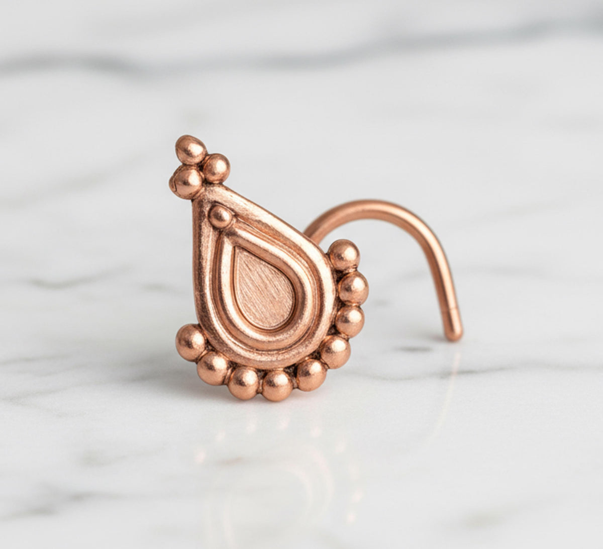 A copper-colored nose ring with a decorative pendant is displayed on a marble surface.