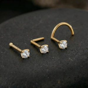 Three gold-colored nose studs with clear gemstones are displayed on a dark surface.