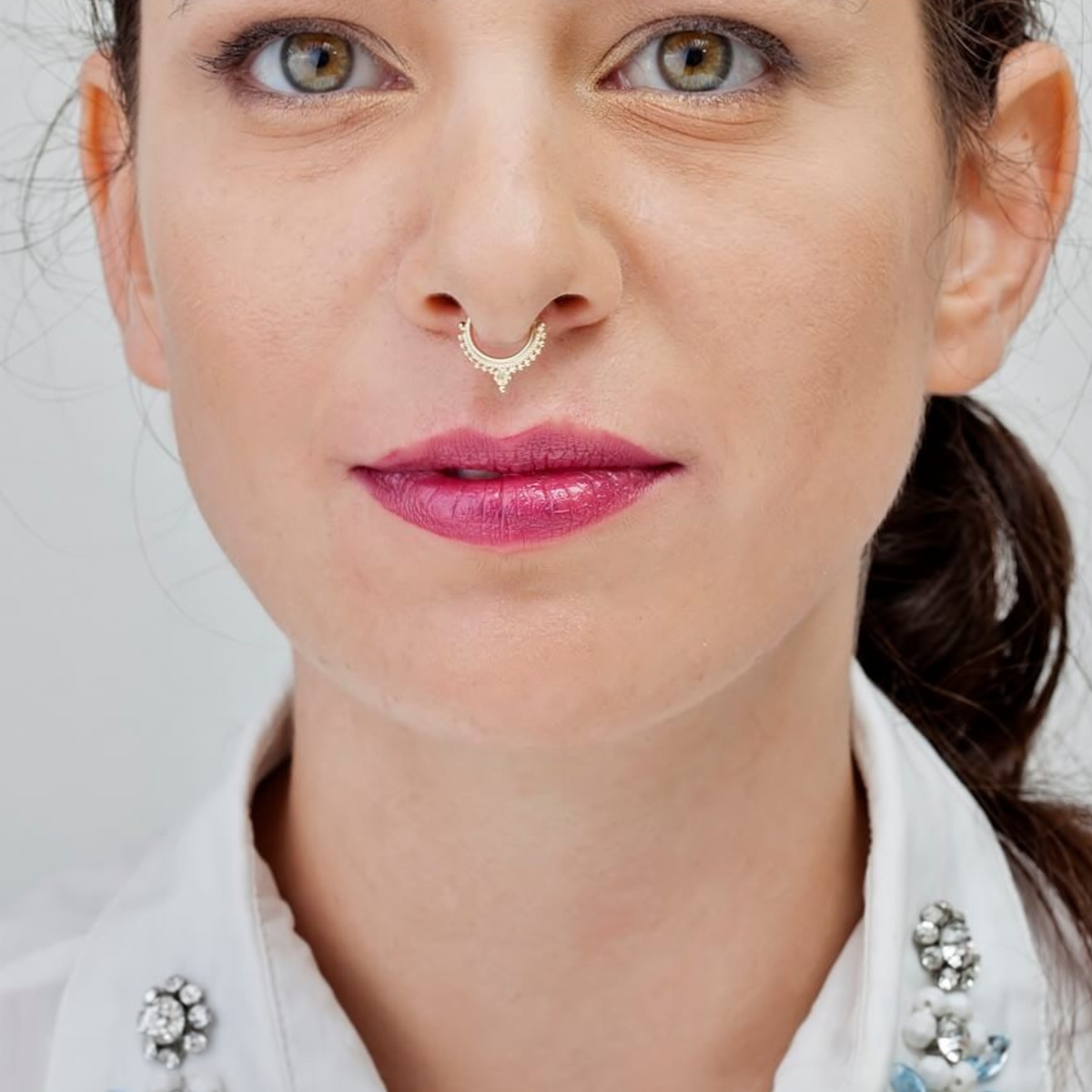 A close-up portrait of a woman with a nose piercing and bold red lipstick, wearing a white collared shirt with a necklace.