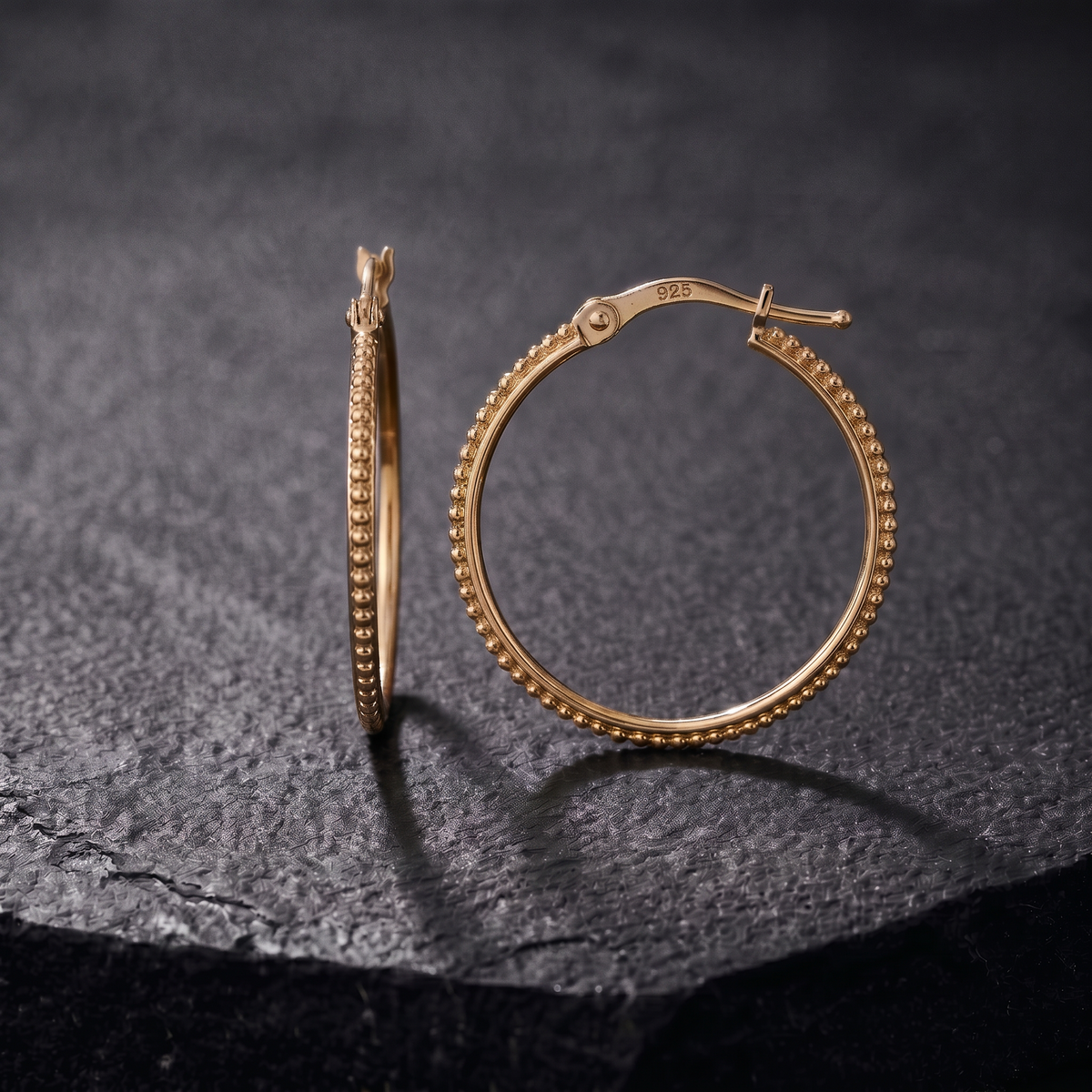 A pair of gold hoop earrings with a textured design, resting on a dark surface.