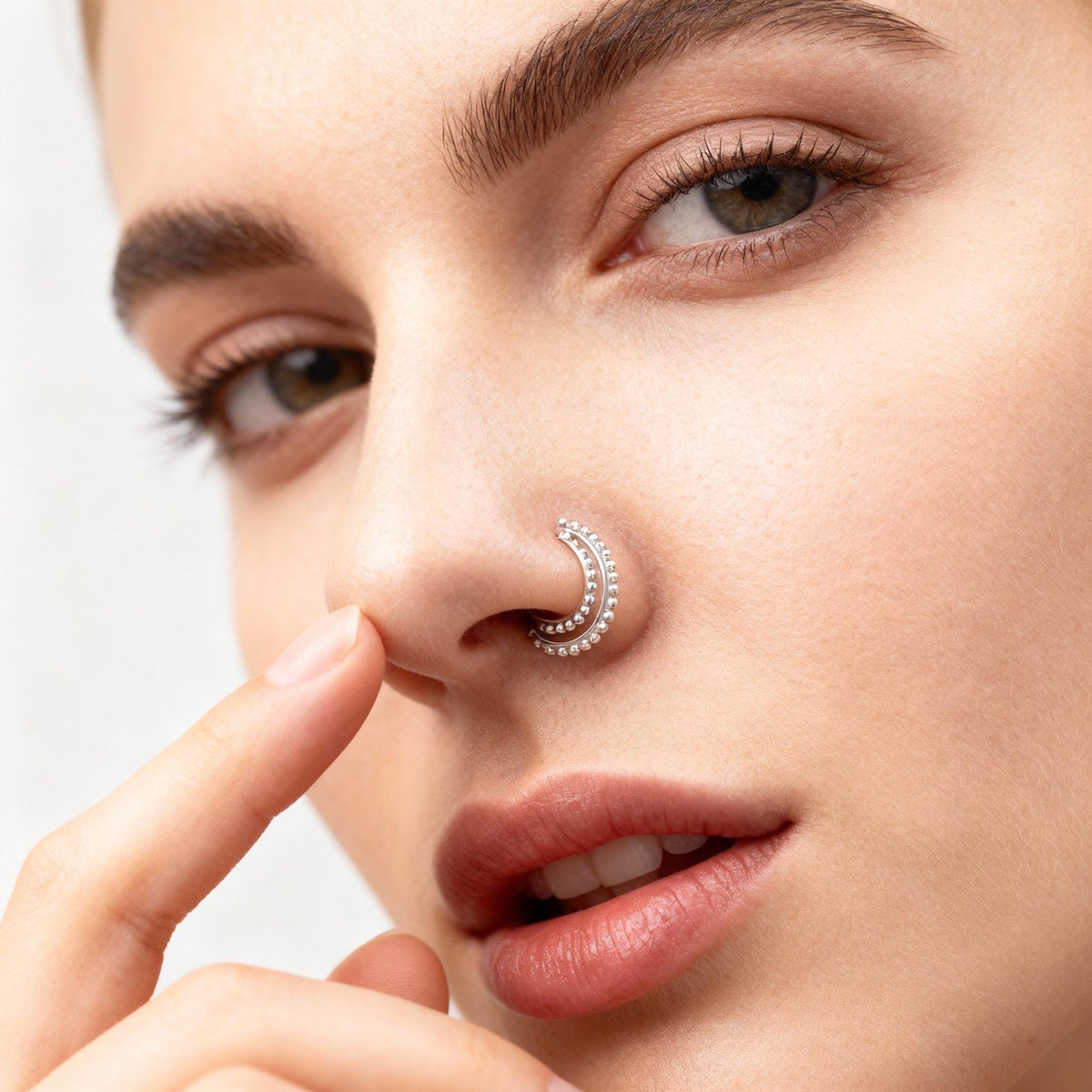 A close-up of a woman's face, focusing on her nose and fingers. She is wearing a nose ring and has her finger on her nose.