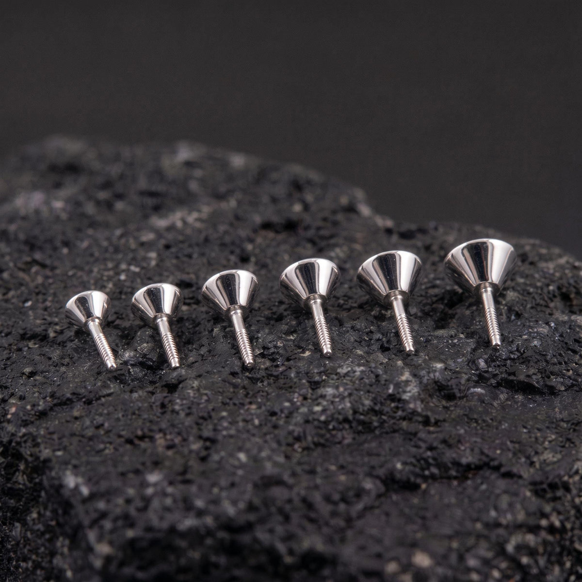 Six silver-colored metal objects are arranged in a row on a dark surface, with their shapes and sizes varying.