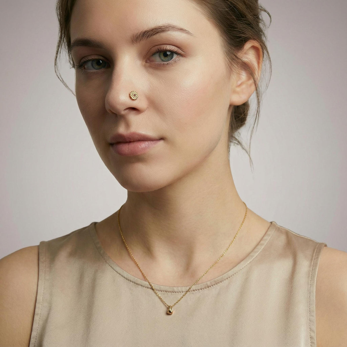 A woman with a nose piercing and a gold necklace is shown in a close-up portrait against a plain background.