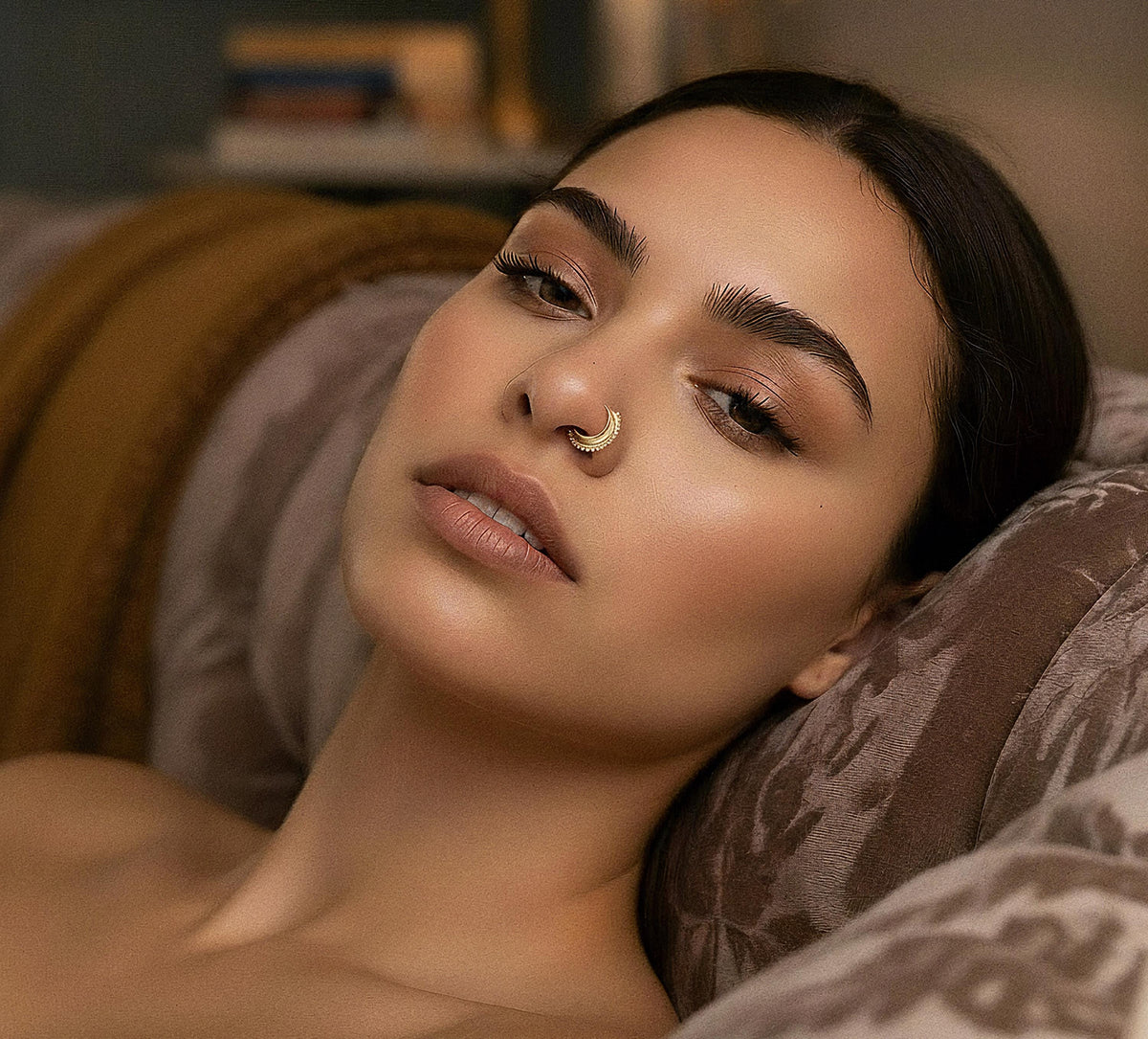 A woman with dark hair and a nose ring is lying on a patterned couch, looking pensive.