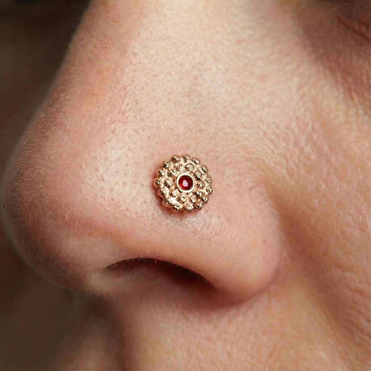 A close-up of a person's nose with a small, ornate gold-colored nose ring with a red gemstone in the center.