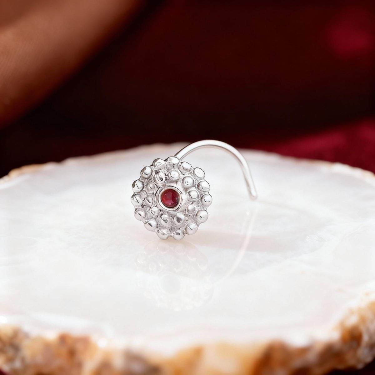 A silver nose ring with a red gemstone is displayed on a white surface against a dark background.