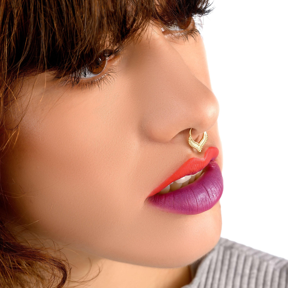 A close-up of a woman's face with long brown hair, wearing a nose ring and vibrant red and purple lipstick.