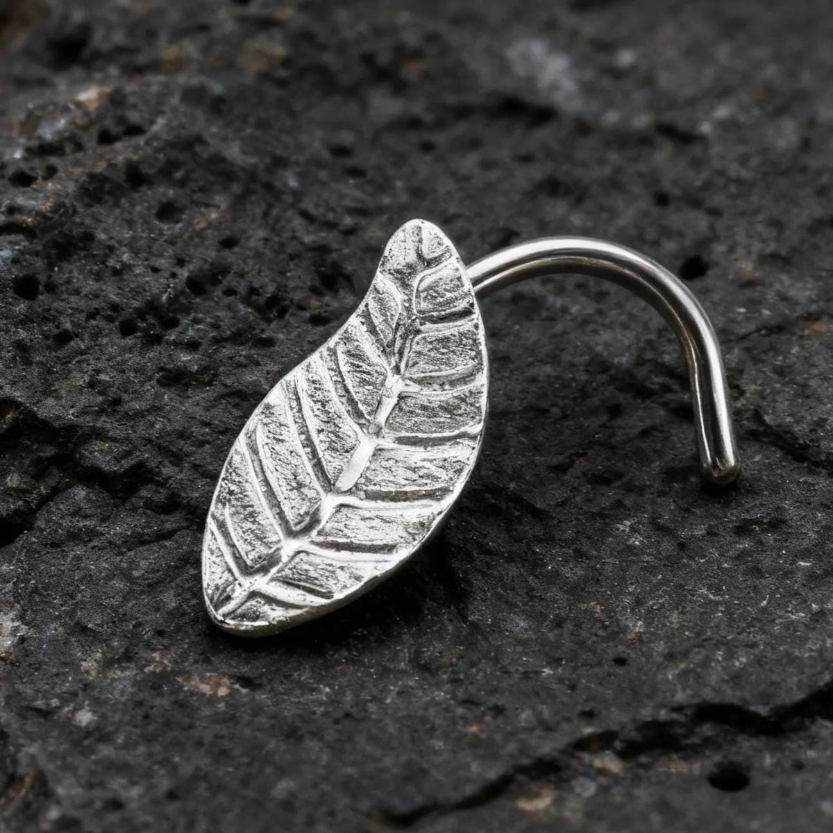 A silver leaf-shaped nose ring is resting on a dark, rocky surface.