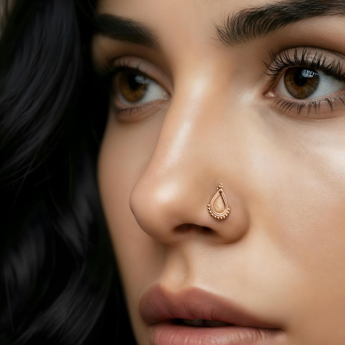 A close-up of a woman's face, focusing on her nose and eyes. She has dark hair and is wearing a gold-colored nose ring.