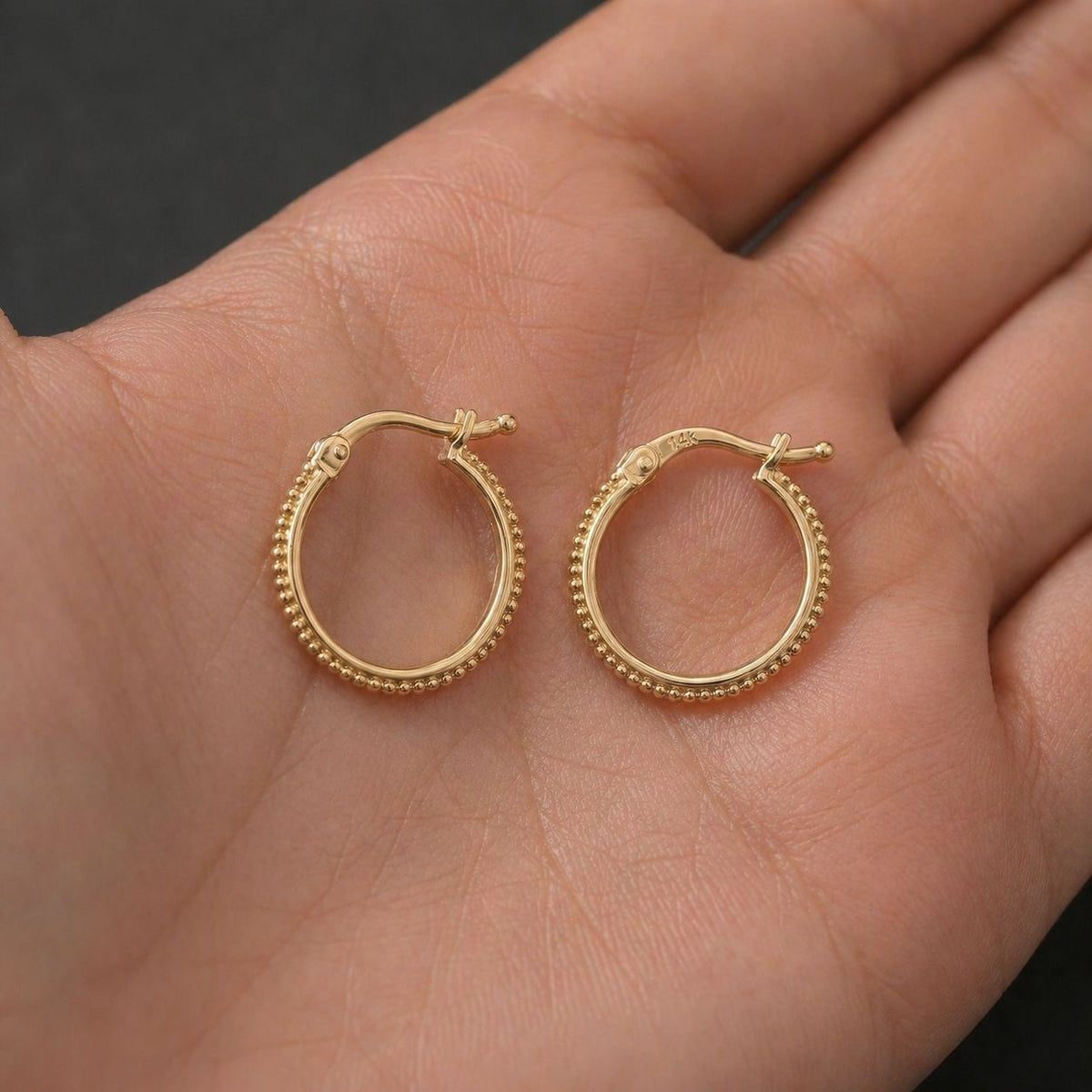 A hand holding two gold hoop earrings against a dark background.