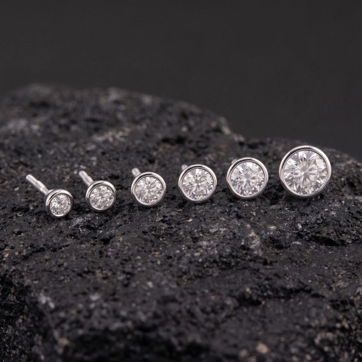 A collection of six diamond-encrusted earrings is displayed on a dark, rocky surface.