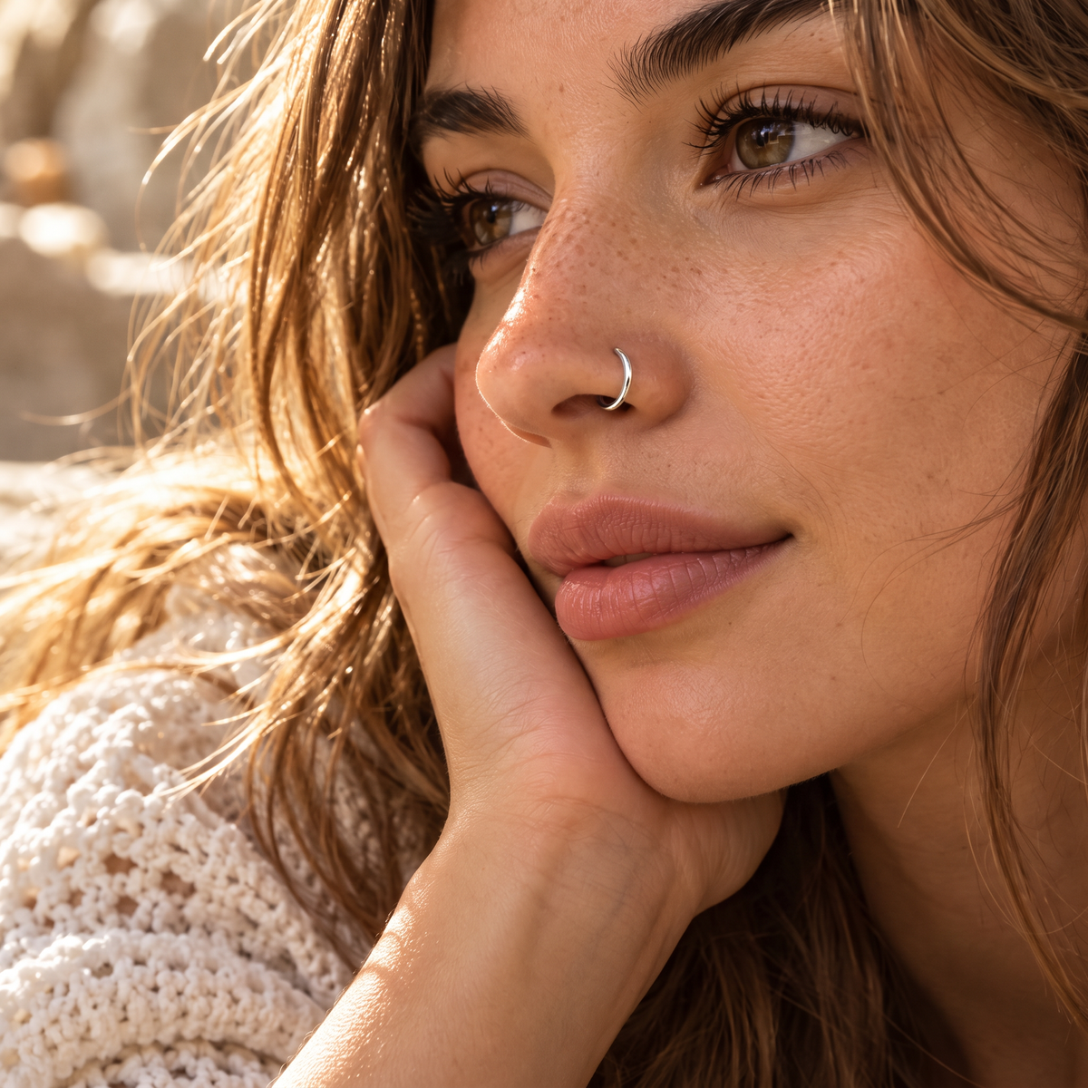 A close-up portrait of a young woman with long, wavy brown hair and a nose ring, looking pensively at the camera.