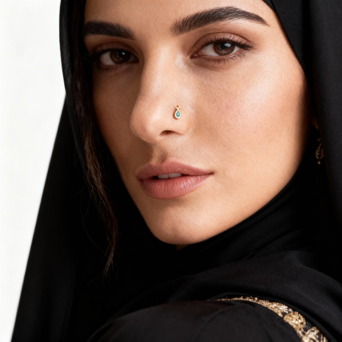 A close-up portrait of a woman wearing a black hijab and a gold-colored nose ring.