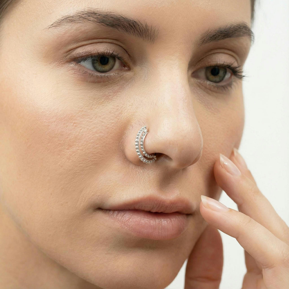 A close-up of a woman's face, focusing on her nose and fingers. She is wearing a nose ring and has her hand resting on her face.
