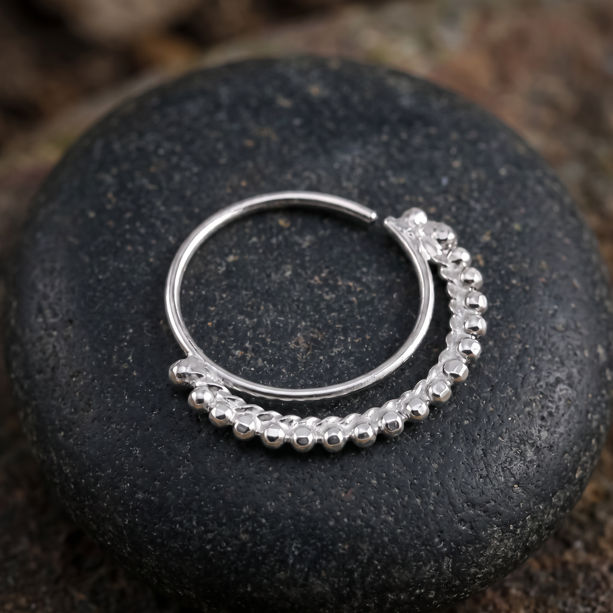 A silver hoop earring with a decorative bead design rests on a dark, textured surface.
