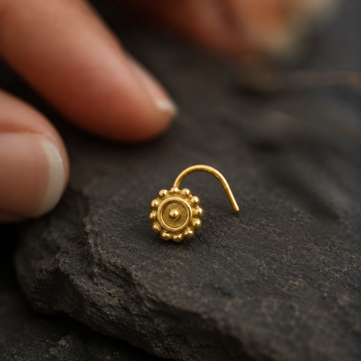 A gold-colored nose stud is resting on a dark surface, with a blurred hand visible in the background.