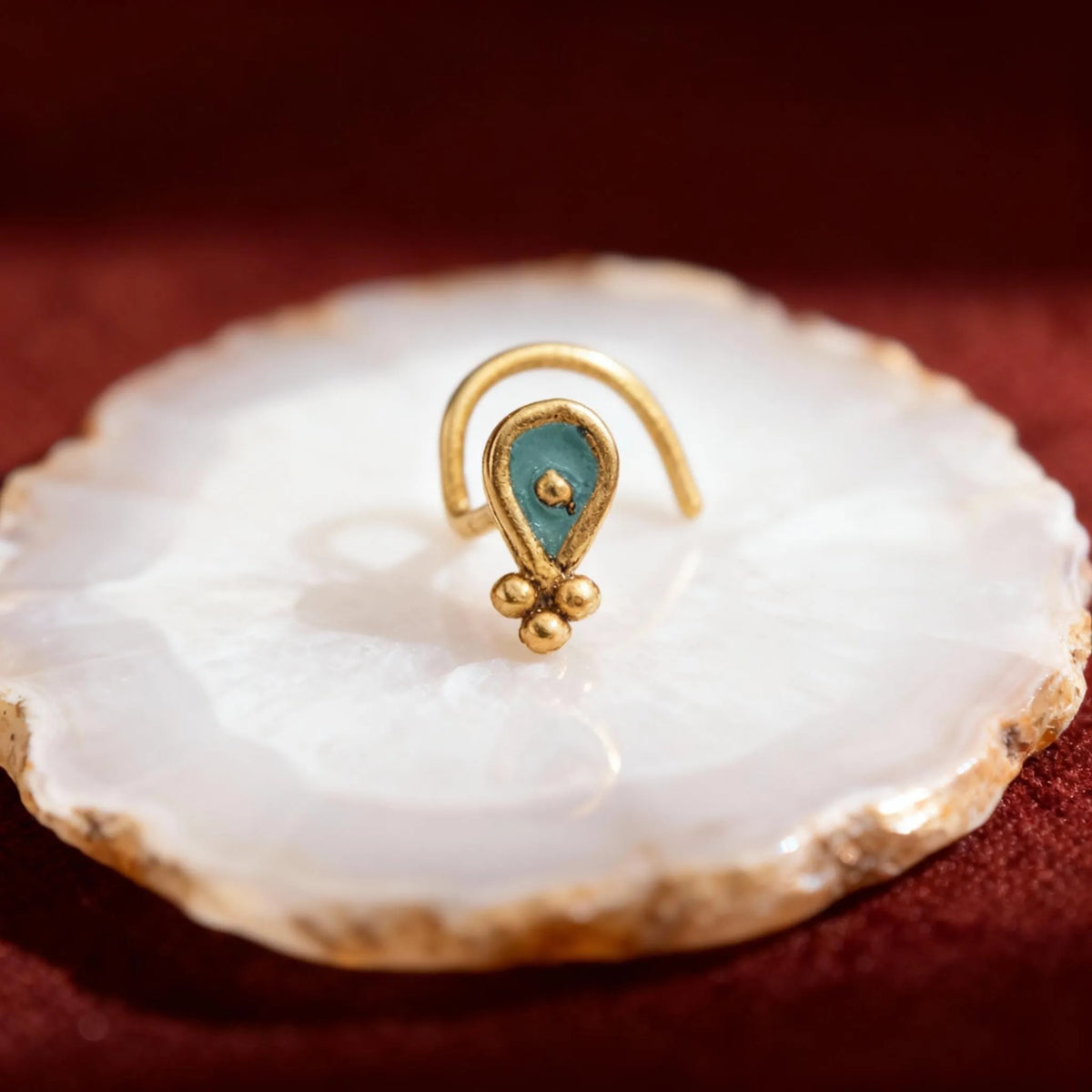 A gold-colored nose ring with a turquoise stone is displayed on a white stone or marble surface against a red background.