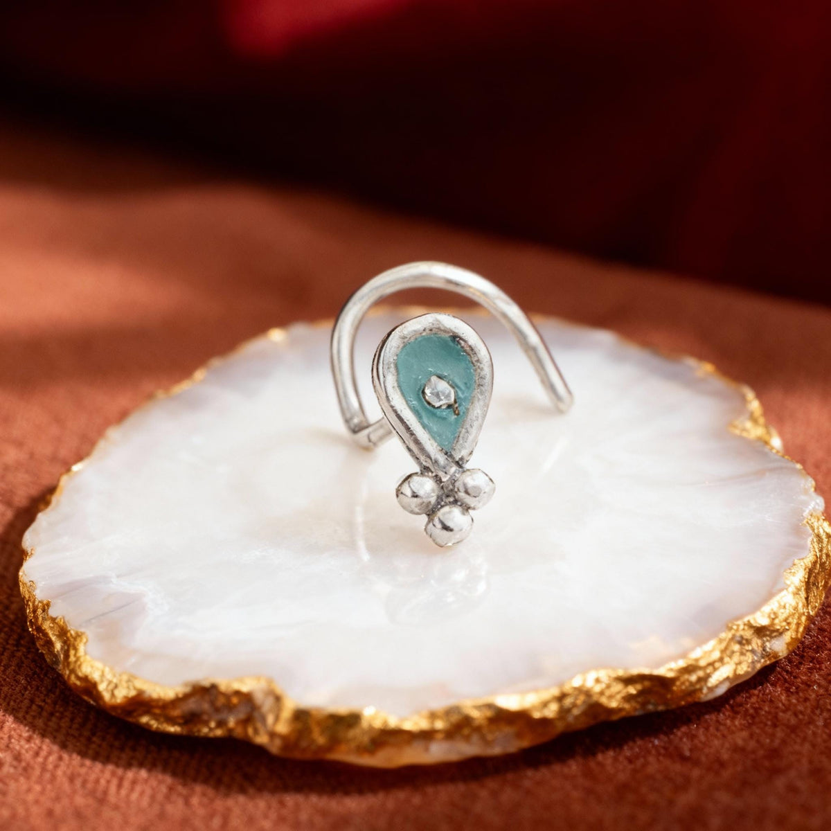 A silver earring with a teal-colored gemstone is displayed on a white and gold stone plate.