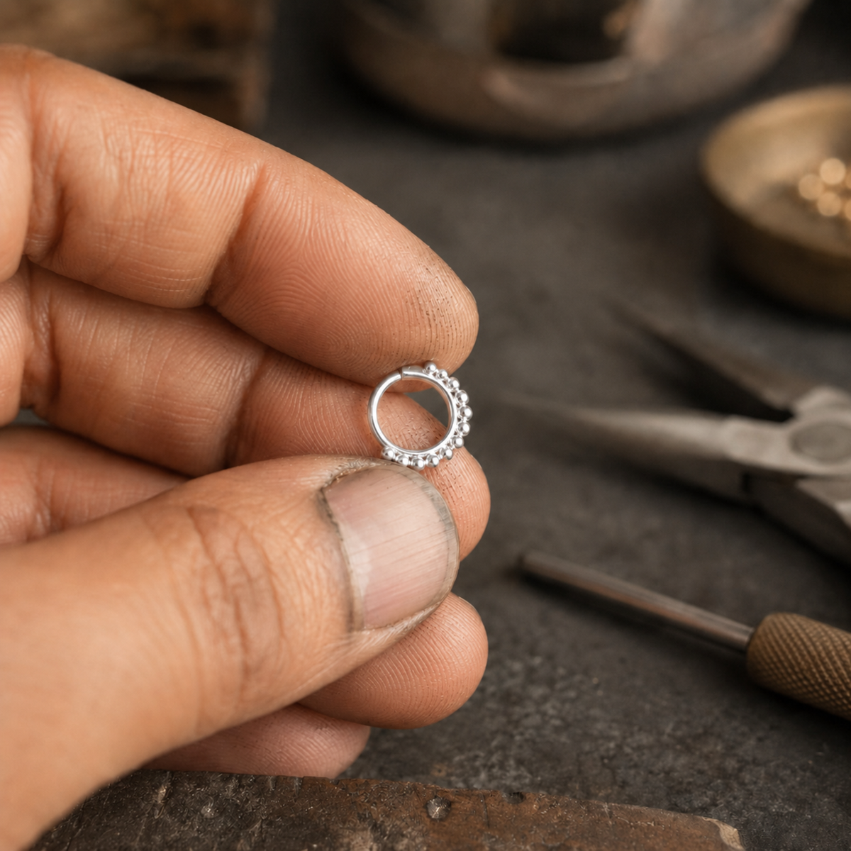 A hand holding a small silver ring with a decorative element on it, surrounded by various jewelry-making tools and materials on a workbench.
