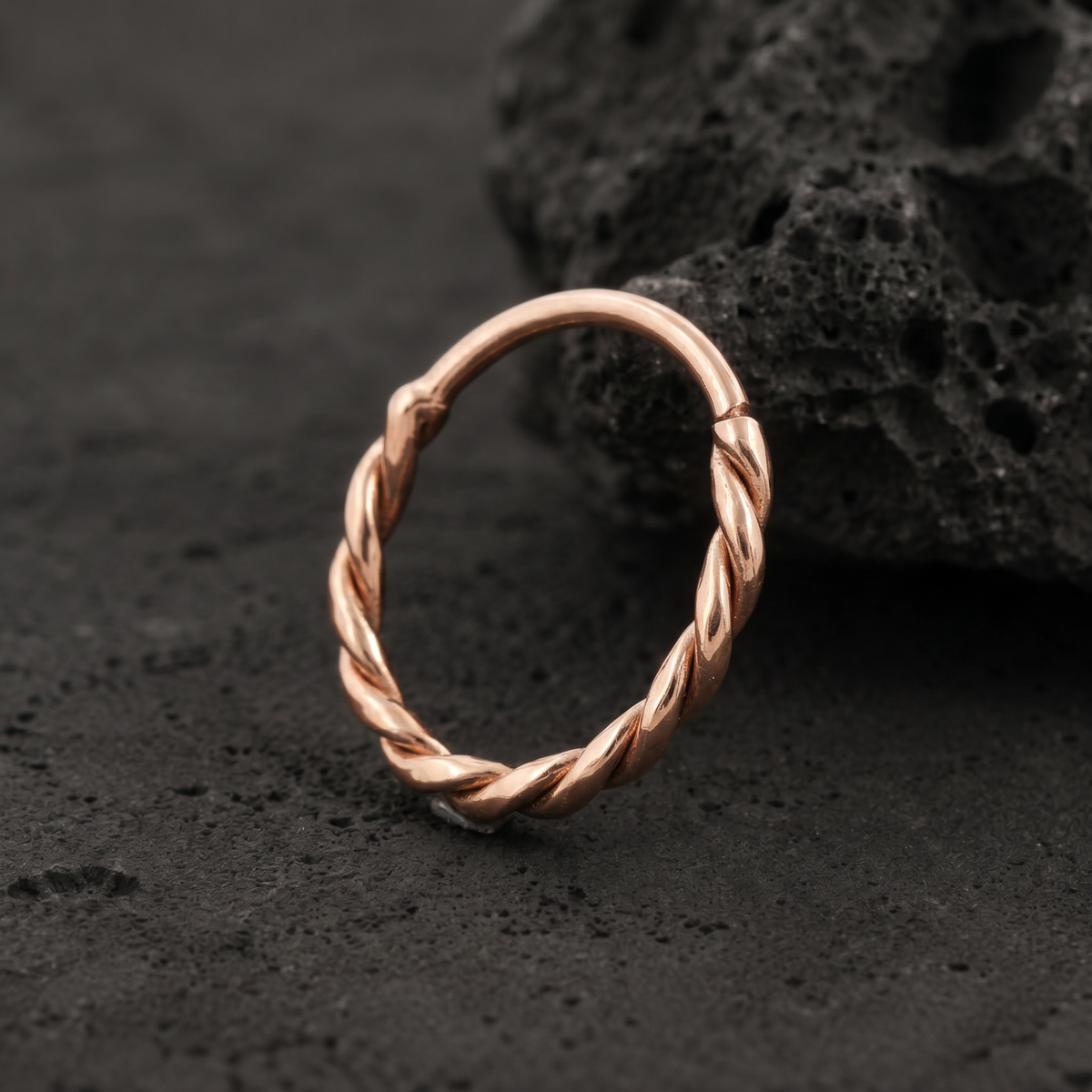 A copper-colored metal ring with a twisted design sits on a dark surface, with a rock-like texture in the background.