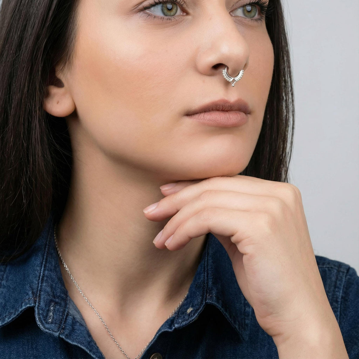 A close-up portrait of a woman with long dark hair, wearing a denim shirt and a nose ring. She is thoughtfully resting her chin on her hand, with a neutral expression on her face.