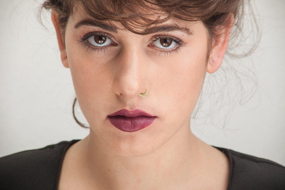 A close-up portrait of a woman with brown hair and a nose piercing, wearing a black top.