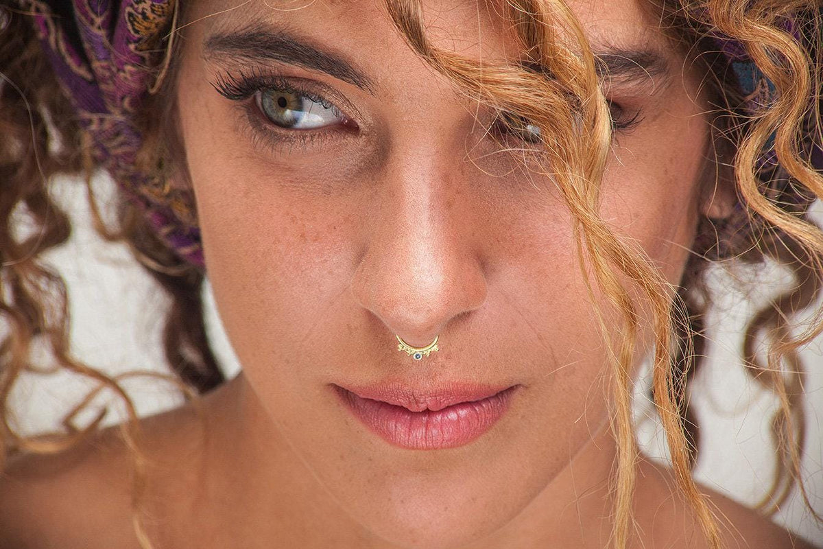 A close-up portrait of a woman with curly hair, wearing a nose ring and looking directly at the camera with a serious expression.